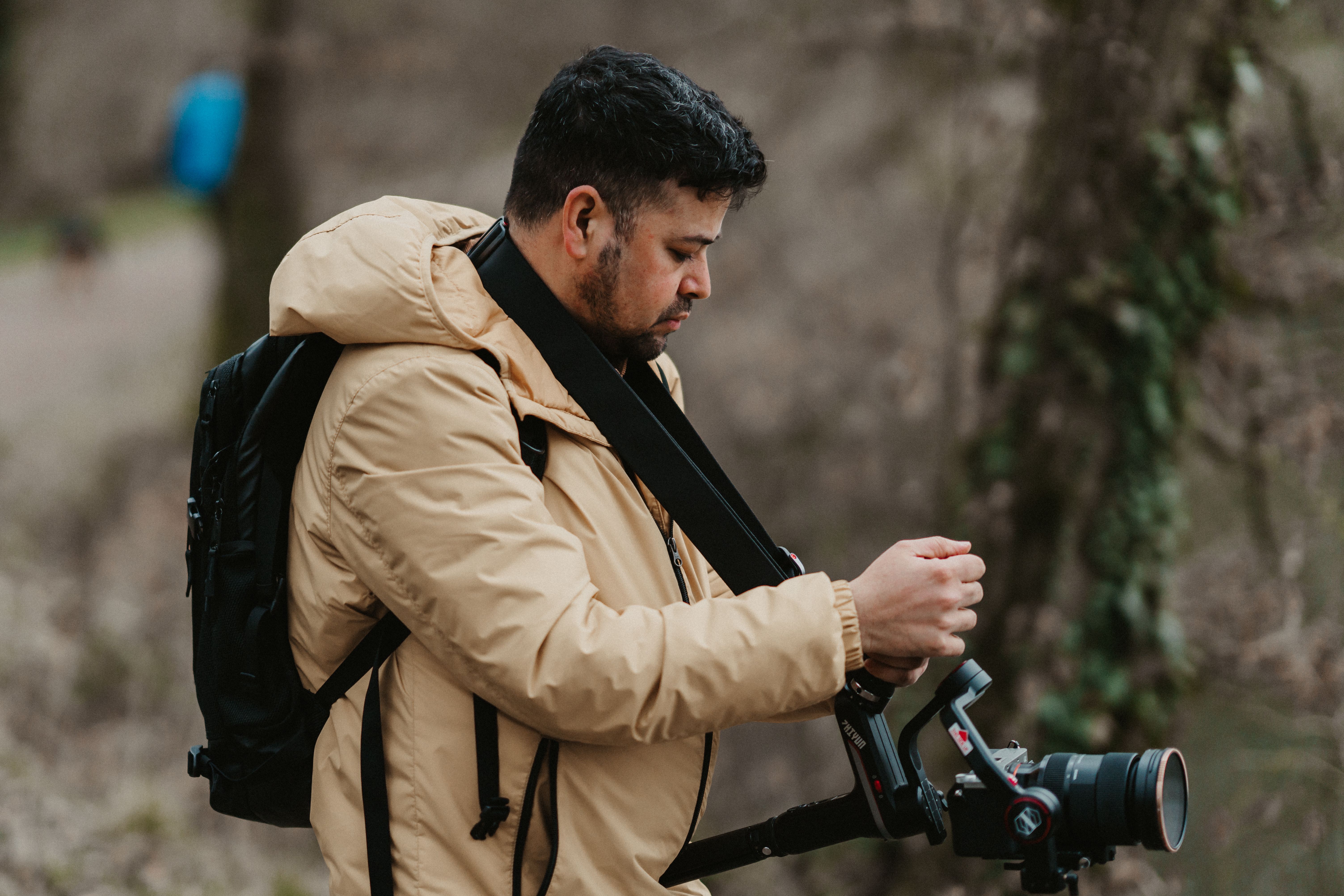 A professional photographer in a beige jacket adjusting his camera in a natural outdoor setting.