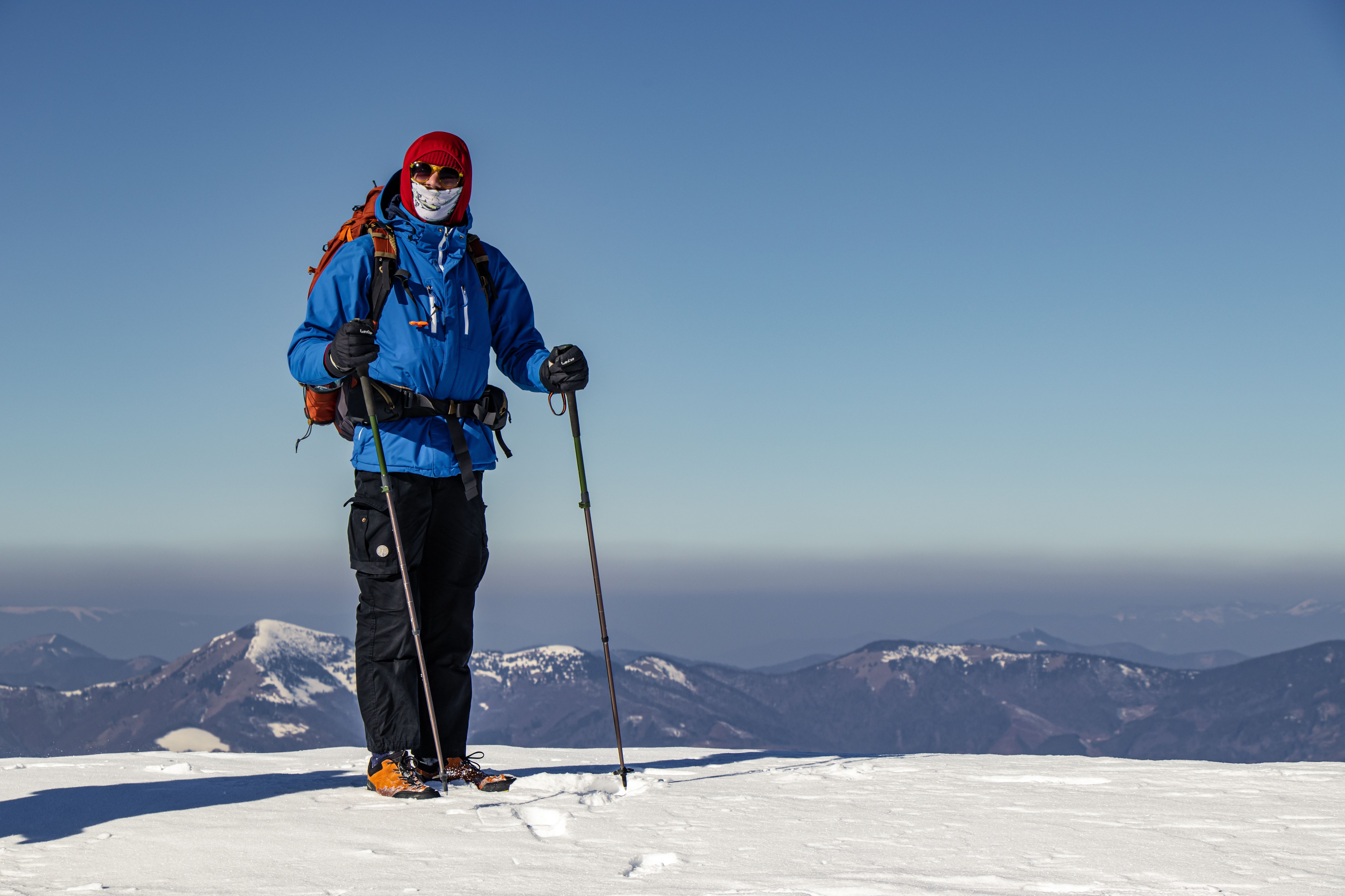 A lone hiker in winter gear stands on a snowy mountain summit with trekking poles.