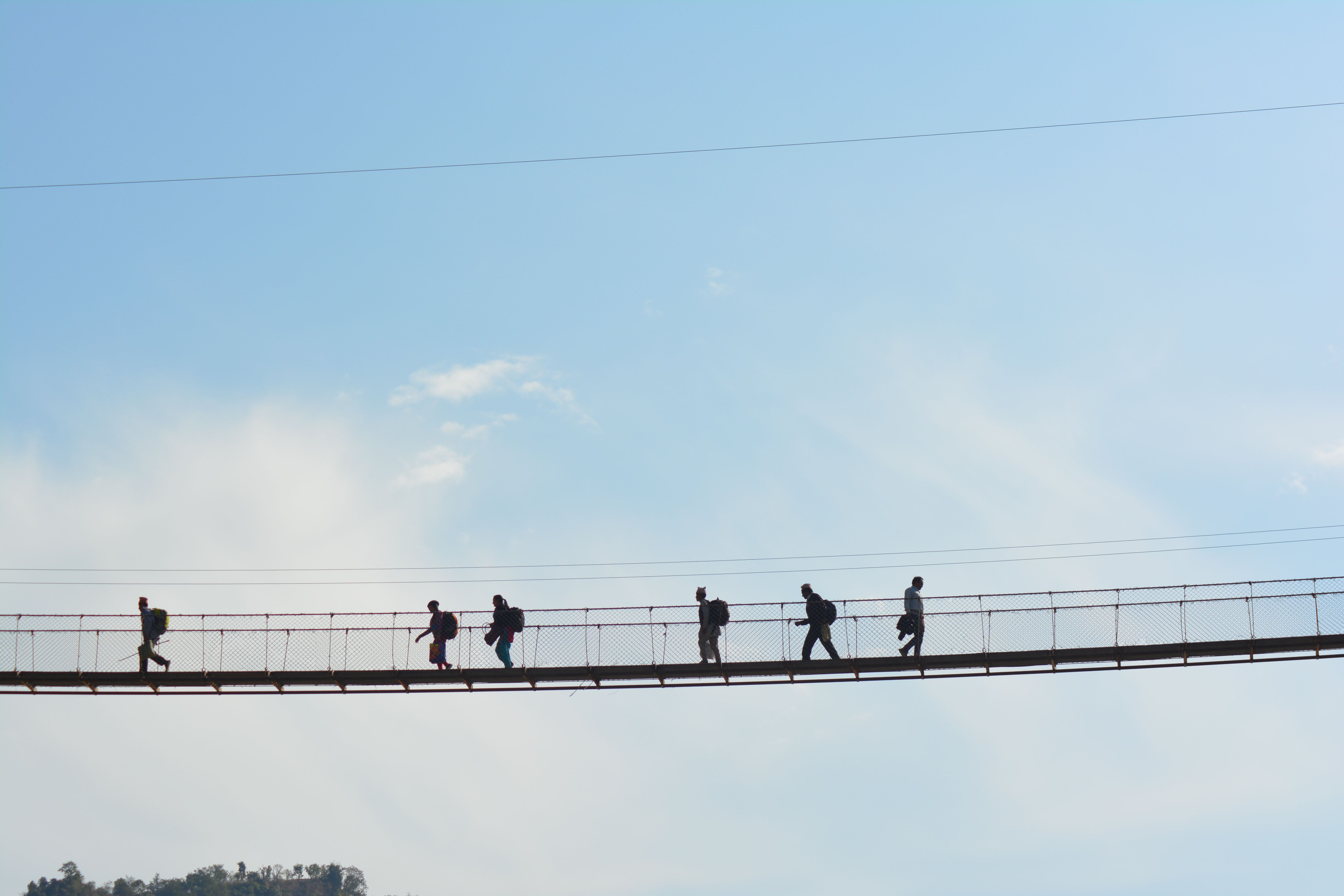 A group of people silhouetted against the sky walking across a suspension bridge during the day.