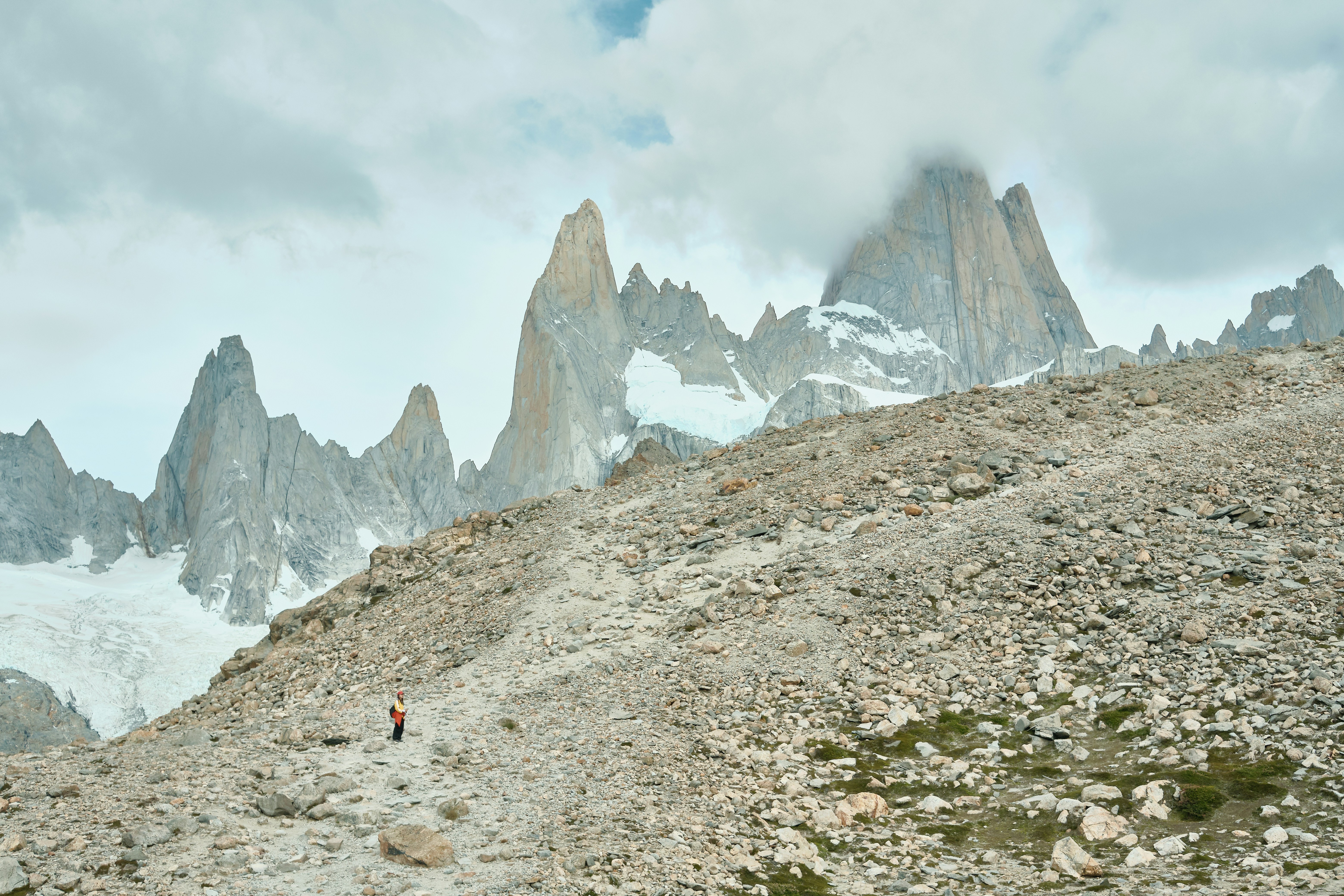 A lone hiker walks towards jagged mountains under cloudy skies.
